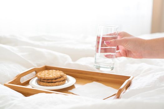 Female hand holding glass of water over tray with crackers on a bed