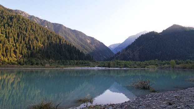 View of the mountain lake Issyk from a height. Turquoise-emerald water from the glacier. Reflection of mountains, forests and the sun on the water. The river flows into the lake. Issyk Dam. Kazakhstan