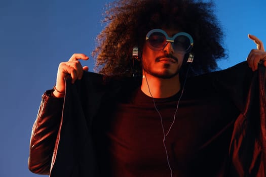 Portrait of a stylish man with curly hair with glasses and headphones on a blue background multinational, colored light, black leather jacket trend, modern concept. High quality photo