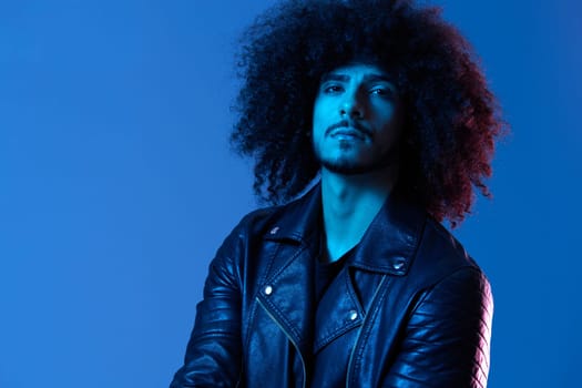 Portrait of fashion man with curly hair on blue background multinational, colored light, black leather jacket trend, modern concept. High quality photo