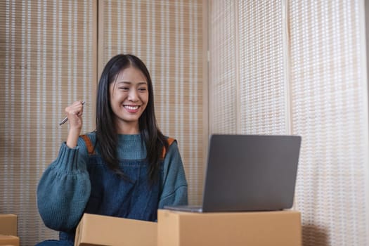 Happy young Asian woman entrepreneur, Smile for sales success after checking order from online shopping store in a laptop at home office, Concept of merchant business online and eCommerce..