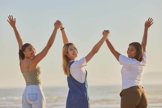 Happy, girl friends and holding hands at a beach with women on a holiday, freedom and travel. Portrait of people with arms raised to show excited adventure by the ocean and sea together by water.
