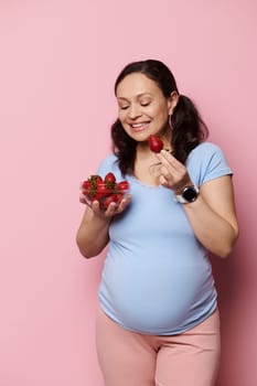 Beautiful multi ethnic pregnant woman holding bowl of sweet ripe organic fresh strawberries, having healthy lunch, isolated pink background. Pregnancy lifestyle. Nutrition. Balanced diet concept