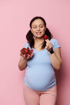 Vertical studio portrait of beautiful vegan pregnant woman, expecting a baby, eating fresh tasty sweet strawberries, isolated pink background. Organic nutrition and balanced diet for expectant mother