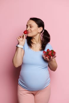 Beautiful glamorous pregnant woman in a blue t-shirt and pink pants, sniffing fresh ripe strawberries, enjoying a healthy diet during pregnancy, posing with bowl of berries on isolated pink background