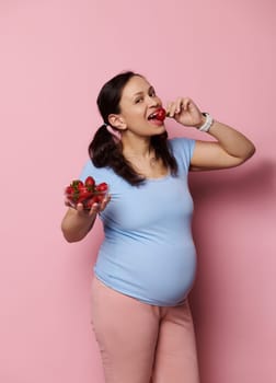 Gorgeous pregnant woman eating fresh organic strawberries, isolated over pink background. Expectant mother in blue t-shirt, holding bowl with fresh berries. Healthy nutrition and pregnancy concept.