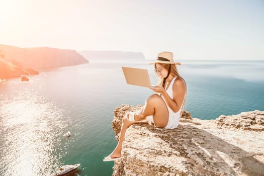 Successful business woman in yellow hat working on laptop by the sea. Pretty lady typing on computer at summer day outdoors. Freelance, travel and holidays concept.