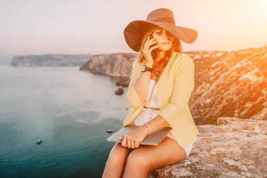 Successful business woman in yellow hat working on laptop by the sea. Pretty lady typing on computer at summer day outdoors. Freelance, travel and holidays concept.