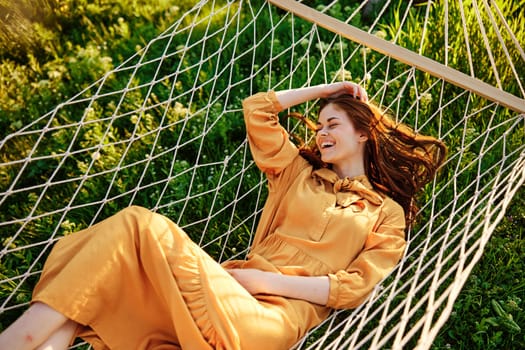 a beautiful, elegant woman lies in a long orange dress on a mesh hammock resting in nature, illuminated by the warm sunset light, happily smiling, straightens her hair with her hands. High quality photo