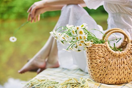 a woman holds a wicker bag with daisies with her hand while sitting on the pier. High quality photo