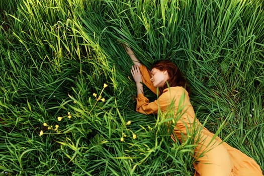 a relaxed woman enjoys summer lying in the tall green grass in a long orange dress stretched out stretching her arm forward. High quality photo