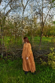 an elegant, sophisticated woman poses relaxed standing near a wicker fence at the dacha in a long orange dress, turning her head to the side. Vertical photography in nature. High quality photo