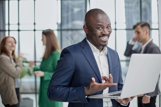 Young African American man in a business suit holding a laptop against the background of his colleagues