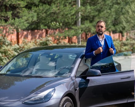 Caucasian bearded man in a blue suit gets out of a black electro car in the countryside in summer