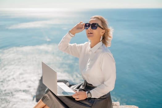 Business woman on nature in white shirt and black skirt. She works with an iPad in the open air with a beautiful view of the sea. The concept of remote work