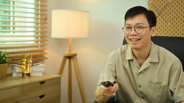 Smiling man holding remote control, watching TV in cozy living room. Entertainment, technology and hobby concept.