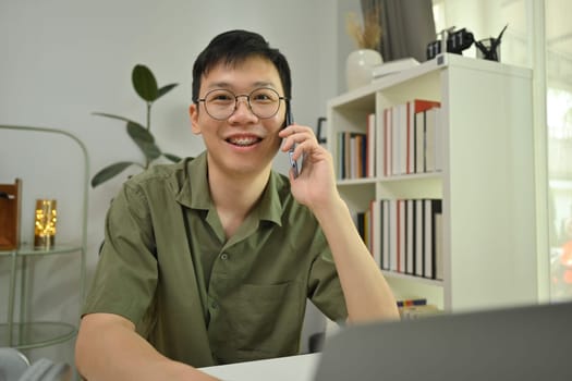 Smiling Asian man freelancer in glasses talking on cellphone and using laptop at desk in home office.
