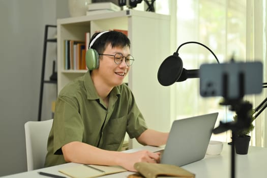 Smiling male podcaster wearing headphone streaming and broadcasting podcast from home studio.