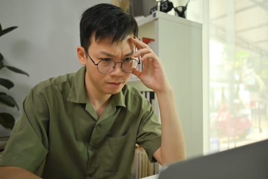 Serious male freelancer in glasses touching forehead while working on laptop at home.