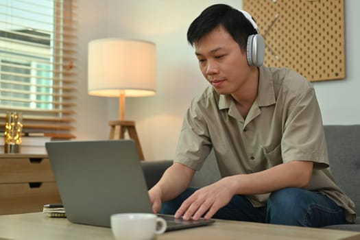 Young Asian man wearing headphone sitting on couch working remotely online on laptop at home.