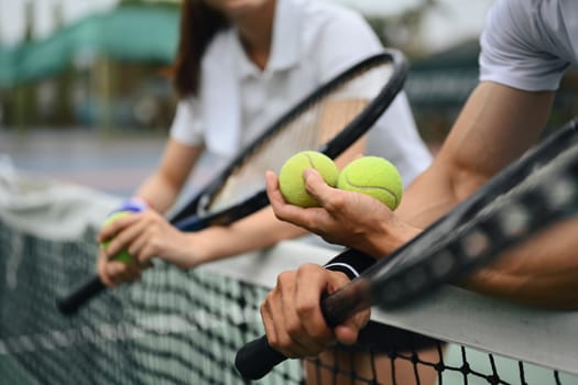 Cropped shot of male tennis coach hand holding balls, giving instructions to his student, standing by net at the court.