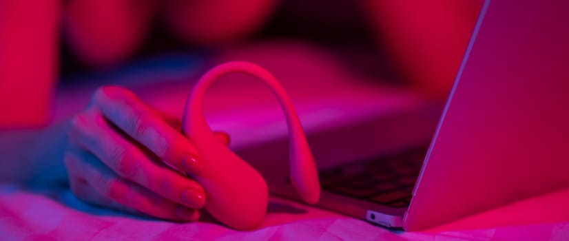 A woman is lying in bed holding a curved vibrator and looking at a laptop. Girl using sex toy in blue-red light