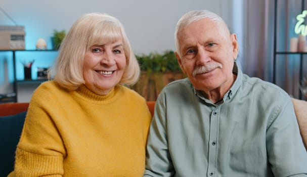 Happy old senior elderly family couple hugging, laughing, smiling looking at camera, healthy retired two grandparents husband wife. Grandmother grandfather embracing sitting on sofa at home, portrait
