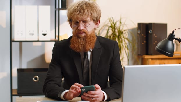 Young businessman in suit use smartphone typing browsing, loses becoming surprised sudden lottery results, bad news, fortune loss fail at home office desk workplace. Bearded freelancer worker man