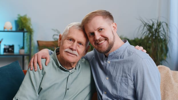 Portrait of senior grandfather with adult man grandson smiling happy embracing hugging. Mature father with young son sitting on couch at home room. Male generations family. Bonding, good relationship