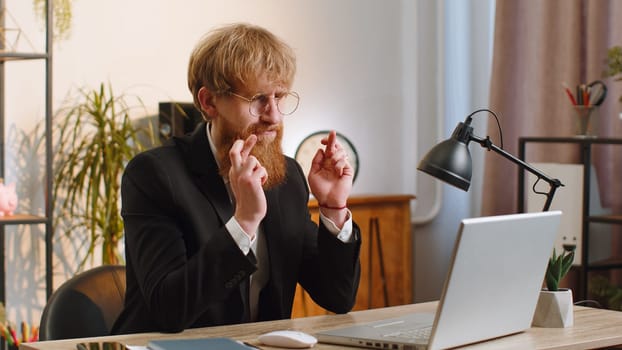Young businessman working on laptop computer at home office desk crossed fingers looking at screen asking for good luck news, wishing good exam results, dreaming about win victory in lottery jackpot