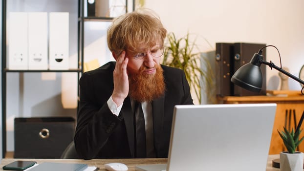 Tired ill young businessman suffering from headache problem tension and migraine, stress at home office desk workplace. Bearded freelancer broker worker man works on laptop notebook. Overworking