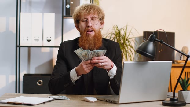 Rich pleased boss businessman guy waving money dollar cash banknotes bills like a fan success business career lottery winner, big income, wealth. Young bearded man freelancer at home office workplace