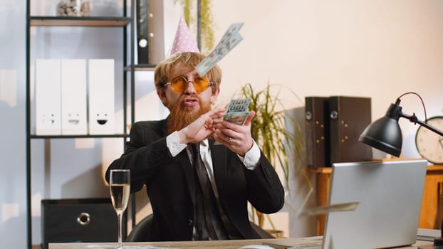 Cheerful rich young businessman working on laptop and wasting throwing money to camera more tips earnings big profit success win lottery shopping. Happy freelancer bearded man at home office desk