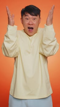 Excited amazed asian man touching head and showing explosion, looking worried and shocked, celebrating success idea. Looking surprised of win wow chinese guy on orange studio background. Vertical shot