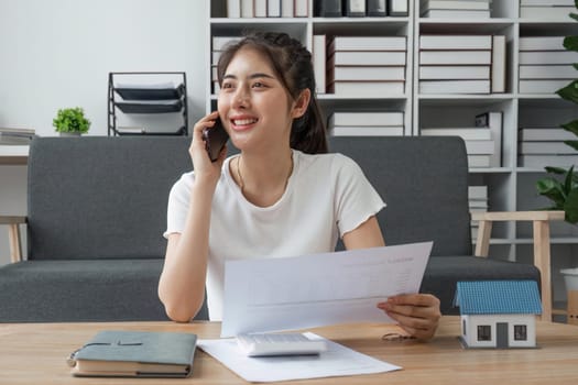 Portrait of smiling young Asian female working remotely, holding paper and reading financial document, talking on mobile phone siting at desk at home office.