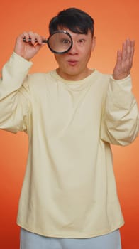 Investigator researcher scientist man holding magnifying glass near face, looking into camera with big zoomed funny eyes, searching, analysing. Asian guy isolated on orange background. Vertical