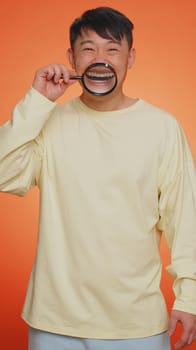 Asian man holding magnifier glass on healthy white teeth, looking at camera with happy expression, showing funny silly face smiling mouth. Chinese guy isolated on orange background. Vertical