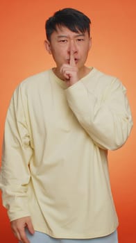 Keep my secret. Do not tell anyone. Shh be quiet please. Asian man presses index finger to lips makes silence gesture sign. Guy on orange studio background. People lifestyles. Vertical shot