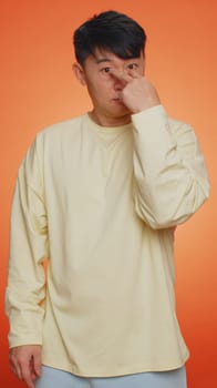 I am watching you. Asian man pointing at his eyes and camera, show I am watching you gesture, spying on someone. Guy isolated on orange studio background. People lifestyles. Vertical view shot