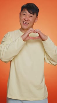 Man in love. Smiling asian man makes heart gesture demonstrates love sign expresses good feelings and sympathy. Handsome chinese adult guy isolated alone on orange studio background. Vertical
