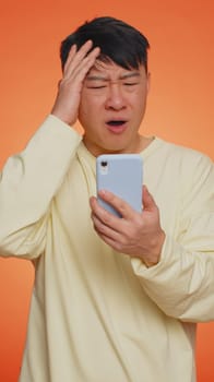 Asian man use mobile cell phone typing browsing, loses becoming surprised sudden lottery results, bad fortune, loss, fail. Handsome chinese guy isolated on orange studio wall background, vertical shot