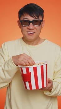 Excited asian man in 3D glasses eating popcorn, watching interesting TV serial, sport game, film, online social media movie content. Guy enjoying domestic entertainment on orange background. Vertical