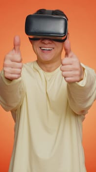 Asian man using headset helmet to play simulation game app online. Watching virtual reality 3D 360 video content. Guy in VR goggles isolated on orange background. Futuristic technology. Vertical shot