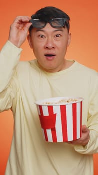 Excited asian man in 3D glasses eating popcorn, watching interesting TV serial, sport game, film, online social media movie content. Guy enjoying domestic entertainment on orange background. Vertical