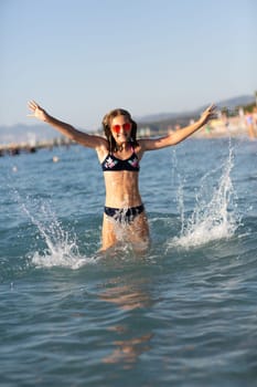 Teenage girl in pink sunglasses having fun on the beach in the sea, playing with waves and splashes. Fun on summer hloiday concept.