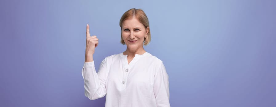 pretty blonde 50s woman in white blouse pointing her finger to the side on studio background with copyspace.