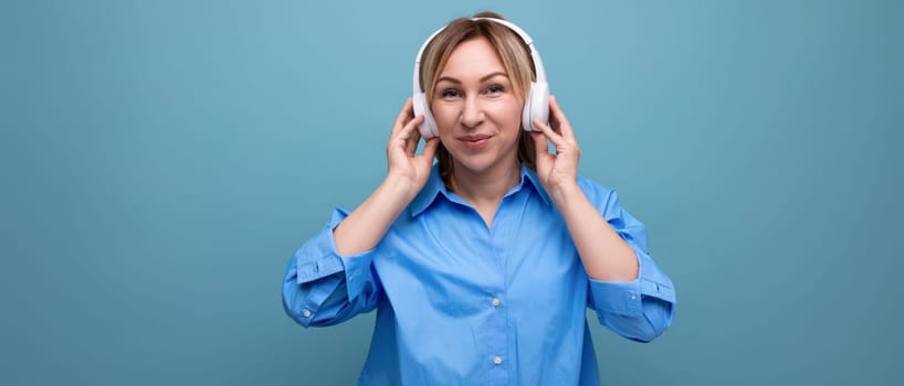 horizontal photo of a smiling young woman in a casual shirt listening to music in big white headphones on a blue isolated background with copy space.