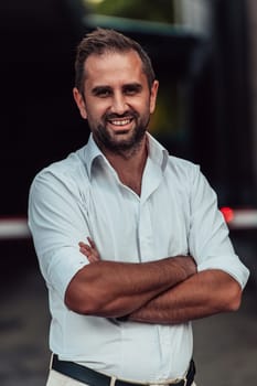 A successful businessman in a white shirt, with crossed arms, poses outdoors, confident expression on his face