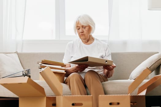 elderly woman sits on a sofa at home with boxes. collecting things with memories albums with photos and photo frames moving to a new place cleaning things and a happy smile. Lifestyle retirement. High quality photo