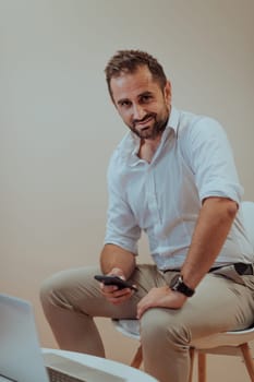 A confident businessman sitting and using laptop and smartphone with a determined expression, while a beige background enhances the professional atmosphere, showcasing his productivity and expertise